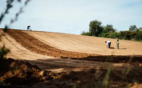 Des ouvriers travaillent sur un terrain en pente, utilisant des outils pour aplanir le sol. L'un mesure avec un mètre, l'autre pelle.