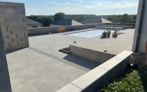 Vaste terrasse en béton gris avec piscine couverte, entourée de végétation et de maisons. Un chantier en cours.
