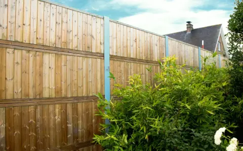 Clôture anti bruit en bois, haute et solide, protégeant un jardin verdoyant. Le ciel bleu et les nuages ajoutent une touche de sérénité.