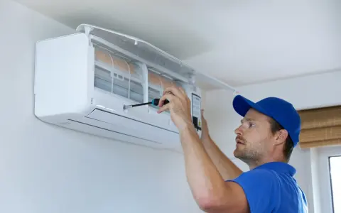 Un technicien en bleu utilise un tournevis pour ajuster un climatiseur mural, assurant le bon fonctionnement pour recharger clim maison.
