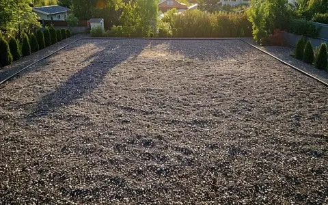Un grand parking en gravier, bordé de haies et d'arbres, sous la lumière dorée du soleil couchant.