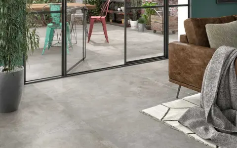 Un salon moderne avec un canapé en cuir marron et un plaid gris. Le sol est recouvert d'un carrelage usé que faire ? Une grande baie vitrée donne sur un patio avec des chaises colorées.