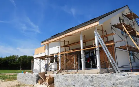 Une maison en construction, avec des échafaudages et une échelle, en cours d'agrandissement. Le ciel bleu est parsemé de nuages légers.