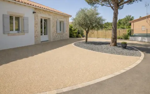 Allée de maison avec béton désactivé de belle épaisseur, bordée de pierres et de gravier sombre autour d'un arbre.