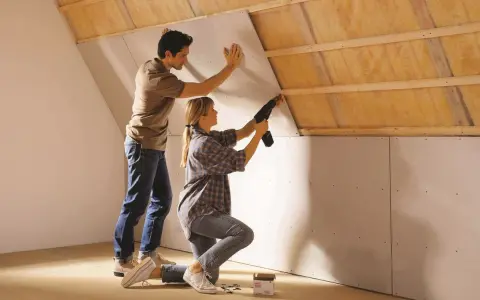 Un couple installe des plaques de plâtre sur un mur mansardé, préparant la surface avant un enduit à la chaux.