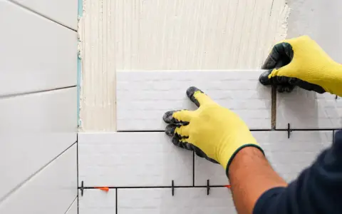 Pose-t-on coller du carrelage avec du ciment ? Oui, un carreleur pose du carrelage blanc sur un mur avec des croisillons.