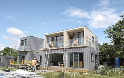 Maison en container prix : construction moderne à deux étages avec balcons, fenêtres panoramiques et un ouvrier en gilet de sécurité.