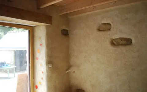 Un mur avec un enduit chaux finition texturé, intégrant des pierres apparentes et des touches de couleur. Le bois des poutres et du cadre de fenêtre apporte de la chaleur.