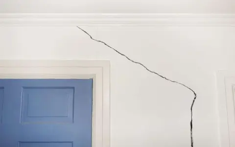 Grosse fissure sur mur intérieur blanc, près d'une porte bleue. Il faut réparer cette fissure pour un intérieur impeccable.