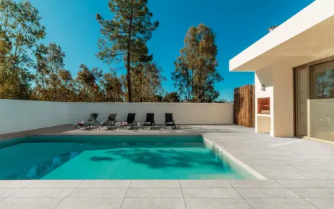 Une piscine moderne avec un sol en carrelage gris clair, des chaises longues et un barbecue extérieur sous un ciel bleu éclatant.