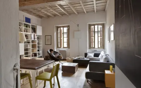 Salon avec poutres apparentes au plafond, une personne lisant dans un fauteuil, canapé, table et étagères.