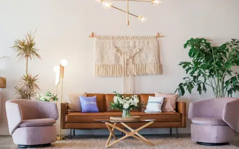Salon moderne avec canapé en cuir marron, fauteuils roses poudrés, tapis texturé et macramé mural. La couleur peinture salon est neutre, mettant en valeur les meubles.