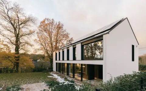 Agrandissement maison ossature bois moderne avec panneaux solaires, entourée d'arbres aux couleurs d'automne et d'un jardin enneigé.