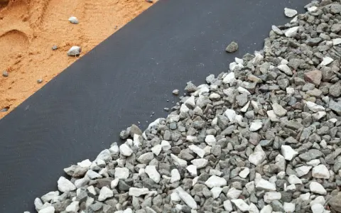 Gros plan sur un géotextile noir recouvert de gravier gris et blanc, à côté de sable. Idéal pour estimer le prix pose géotextile et gravier.