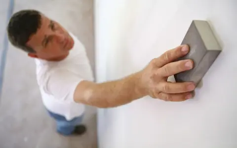 Un homme ponce un mur blanc avec une cale à poncer, préparant la surface avant la peinture.