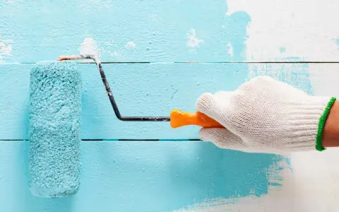 Main tenant une main gantée, un rouleau applique une peinture bleu turquoise sur un mur en bois blanc.