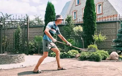 Un homme en chapeau et short utilise un râteau pour faire une place de parking dans son jardin. Le soleil brille sur la pelouse.
