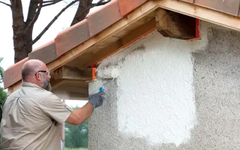 Un homme applique une peinture sur mur béton extérieur avec un rouleau. La rénovation progresse sous le toit de tuiles.