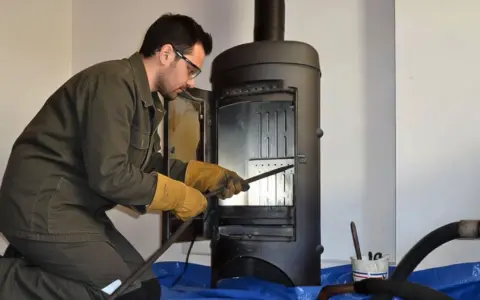 Un homme en tenue de travail effectue le ramonage d'un poêle à bois, ganté et muni d'outils.
