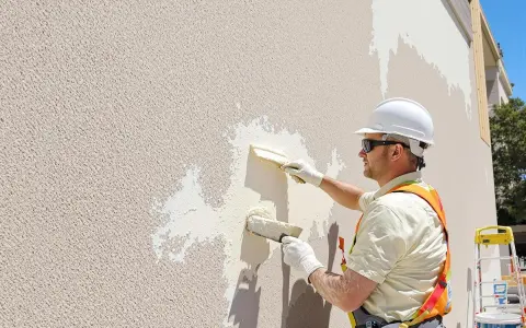 Un ouvrier en casque blanc applique un nouvel enduit extérieur sur un enduit existant, à l'aide de deux truelles.