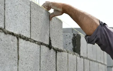 Main d'un maçon plaçant une parpaing pour monter un mur en parpaing de 2m. Le fil à plomb guide la pose.