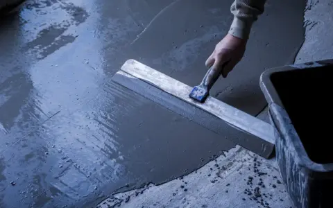 Main d'un ouvrier réalisant un ragréage de terrasse extérieur avec une lisseuse, assurant une pente pour l'évacuation de l'eau.
