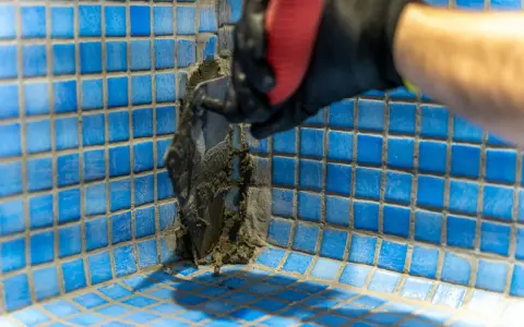 Application de mortier pour l'étanchéité d'une piscine en béton. Un ouvrier en gant noir et rouge applique le produit avec une spatule sur les carreaux bleus.