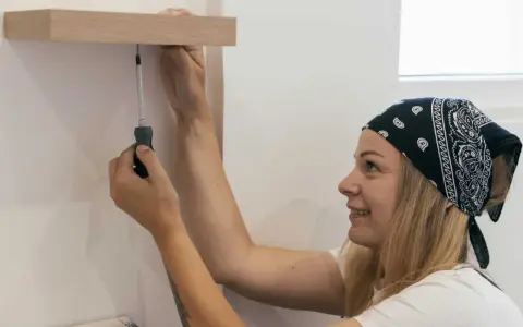 Une femme souriante, bandeau sur la tête, utilise un tournevis pour fixer une étagère en bois. Elle sait comment percer droit pour un résultat parfait.