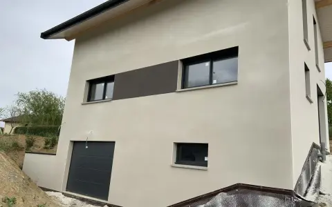 Maison moderne avec enduit neuf, une trace blanche discrète sur le crépi clair. Fenêtres noires et garage gris.