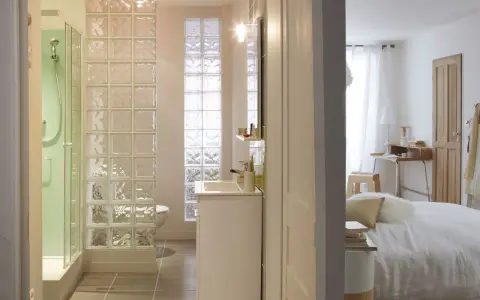 Salle de bain moderne avec douche, lavabo et toilettes, séparée de la chambre par un mur en brique de verre.