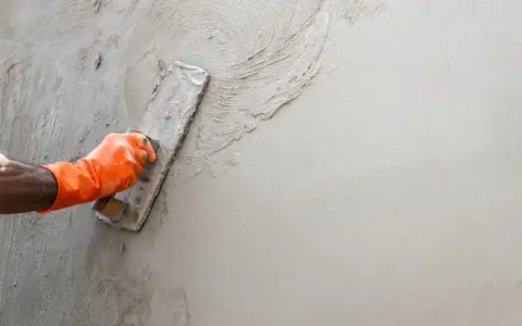 Main d'un ouvrier en gant orange appliquant de l'enduit sur un mur avec une truelle. Apprentissage de comment faire un enduit sur un mur.