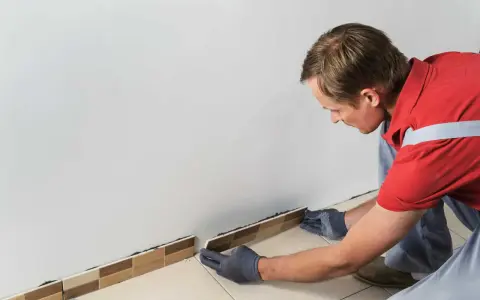 Un homme en gants gris est en train d'enlever la plinthe de carrelage le long d'un mur blanc.