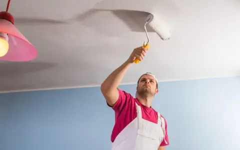 Un homme en salopette blanche et t-shirt rose utilise un rouleau pour peindre un plafond. Il est concentré sur sa tâche, montrant comment peindre un plafond.
