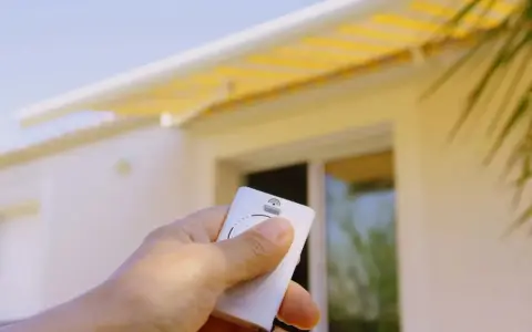 Main tenant une télécommande pour le réglage store banne manuel. Le store jaune est déployé sur une maison blanche.