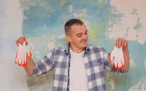 Homme souriant tenant des gants blancs à bouts orange devant un mur texturé. Il pense au prix peinture m2 au black pour son projet.
