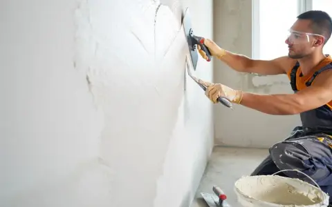 Un ouvrier applique un enduit sur un mur humide avec une truelle et une taloche. Le travail progresse, laissant une surface lisse.