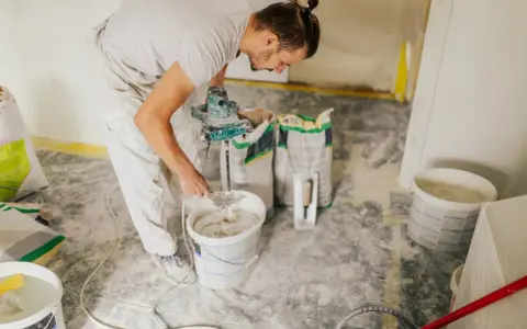 Un ouvrier prépare de l'enduit béton cellulaire dans un seau avec un mélangeur. Des sacs de poudre sont à proximité.