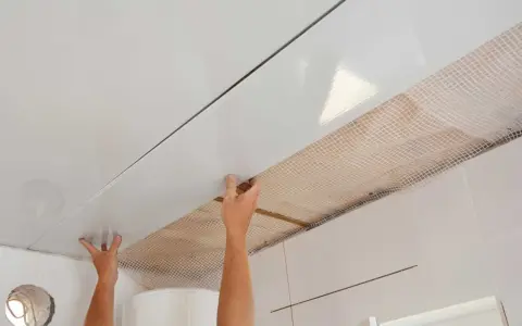 Pose de lambris PVC au plafond avec clips. Les mains ajustent une lame blanche brillante sur une structure, révélant un treillis blanc en dessous.
