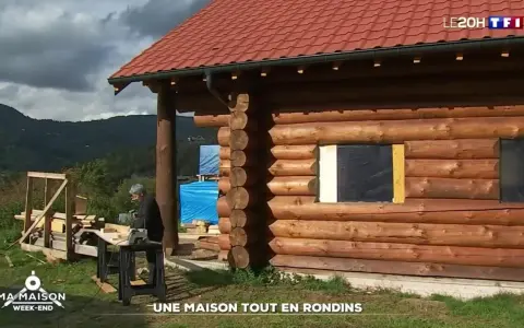 Une maison en rondins de bois en construction, avec un homme travaillant à proximité. Le toit est rouge et le paysage est montagneux.