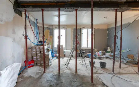 Chantier d'une maison phenix 1970 : poutres métalliques, étais, tuyaux et matériaux de construction dans une pièce en rénovation.
