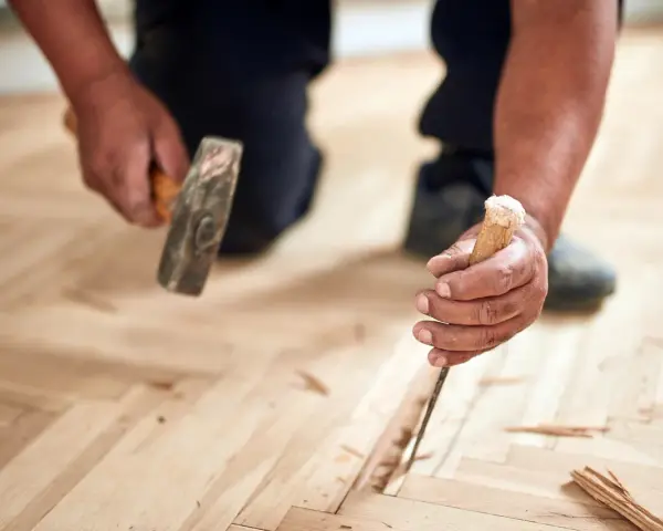 Un artisan utilise un ciseau et un marteau pour changer une lame de parquet flottant endommagée.