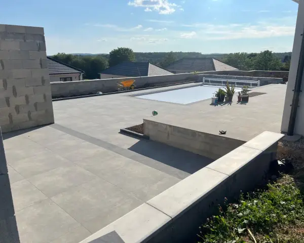 Vaste terrasse en béton gris avec piscine couverte, entourée de végétation et de maisons. Un chantier en cours.