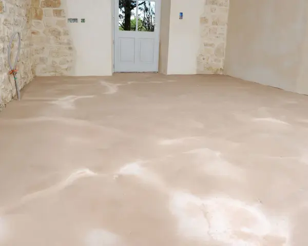 Sol fraîchement ragréé, l'épaisseur du ragréage intérieur est visible sur le sol brut. Les murs en pierre et la porte blanche annoncent une rénovation.