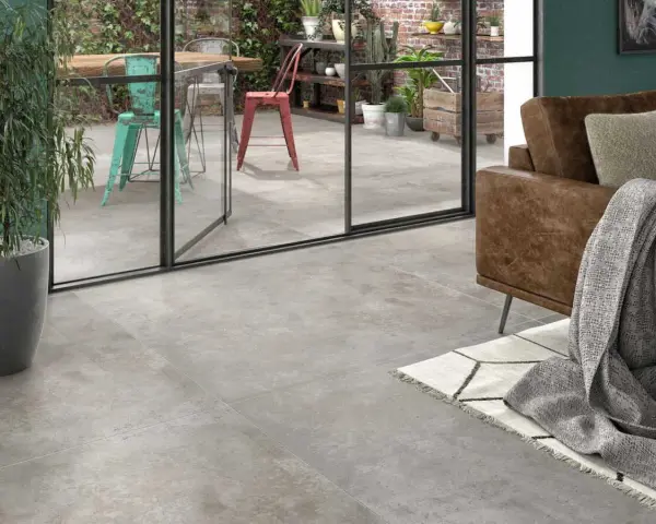 Un salon moderne avec un canapé en cuir marron et un plaid gris. Le sol est recouvert d'un carrelage usé que faire ? Une grande baie vitrée donne sur un patio avec des chaises colorées.