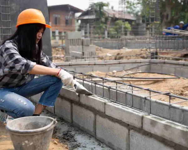 Une ouvrière en casque orange pose du ciment sur un mur en parpaing. Le chantier progresse.