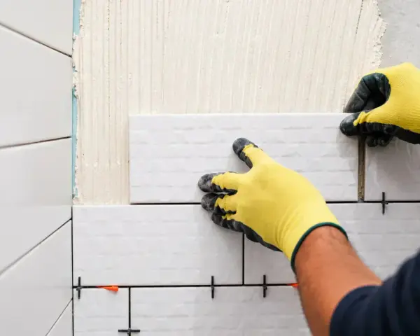 Pose-t-on coller du carrelage avec du ciment ? Oui, un carreleur pose du carrelage blanc sur un mur avec des croisillons.