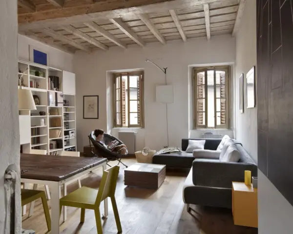 Salon avec poutres apparentes au plafond, une personne lisant dans un fauteuil, canapé, table et étagères.