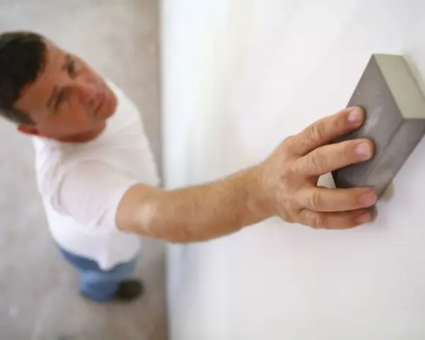 Un homme ponce un mur blanc avec une cale à poncer, préparant la surface avant la peinture.