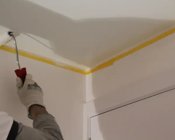 Maintenant, vous savez comment peindre un plafond : une main gantée utilise un rouleau pour peindre le coin d'un plafond, protégé par du ruban de masquage jaune.