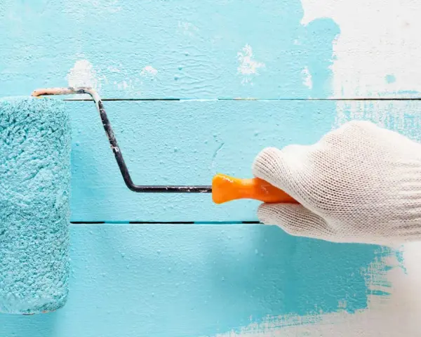 Main tenant une main gantée, un rouleau applique une peinture bleu turquoise sur un mur en bois blanc.